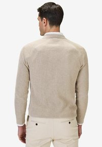 Beige ribbad tröja med långa ärmar, rund halsringning och subtil textur. Har en vit skjortkrage synlig vid halsen.