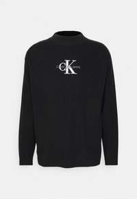 Schwarzer, gerippter Langarmpullover mit hohem Kragen und einem weißen Calvin Klein Jeans-Logo auf der Vorderseite. Weicher Stoff, lockere Passform.