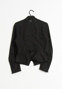 Svart jacka med en strukturerad design, långa ärmar, skräddarsydd midja och detalj med knytning i ryggen. Slät tygfinish med dragkedjor på ärmsluten.