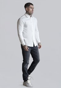 White long-sleeve button-up shirt paired with dark jeans and white sneakers. The shirt features a structured collar; the fabric appears smooth.