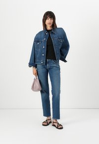 Veste en denim avec une coupe décontractée, de couleur bleue, et poches avant, associée à un jean slim bleu. Des sandales noires et un petit sac à main gris complètent le look.