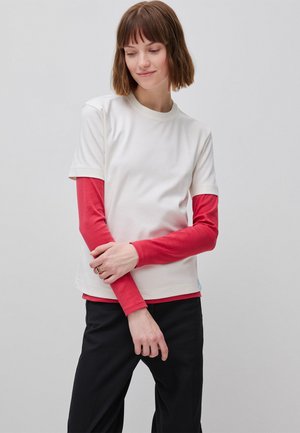 Młoda osoba z krótkimi brązowymi włosami, ubrana w białą koszulkę z krótkim rękawem na czerwonej koszulce z długim rękawem oraz czarne spodnie, stojąca z założonymi rękami.