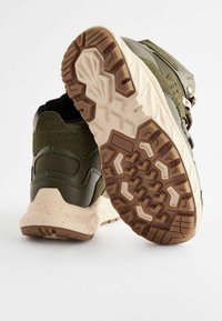Zielone buty sportowe z teksturowaną cholewką, amortyzowaną podeszwą z brązowym bieżnikiem i akcentami w ciemniejszym odcieniu zieleni.