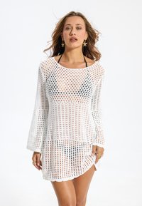 Robe de couverture blanche en crochet à manches longues, présentant un motif à mailles larges et ouvertes avec un ourlet festonné. Convient pour la plage.
