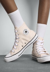 Beige høyde sneakers med kanvasoverdel, hvite hull og snøringer. Har en svart og hvit stjernelogo på siden, gummitåhette og såle.