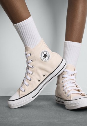 Beige hoge sneakers van canvas met witte veteroogjes en veters. Heeft een zwart-wit sterlogo aan de zijkant, een rubberen teenkap en een zool.
