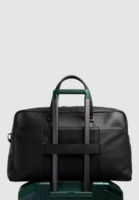 Sac de voyage en cuir noir avec surface texturée. Comprend des poignées supérieures et une sangle pour fixation trolley. Positionné sur une valise verte.