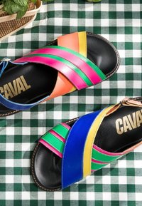 Sandales en cuir colorées avec des brides croisées en rose, vert, bleu, jaune et orange sur un tissu à carreaux vert et blanc.