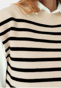Beige pletený svetr se vodorovnými černými pruhy. Klasický kulatý výstřih s žebrovanou texturou. Stylizováno s bílou košilí s límečkem pod ním.