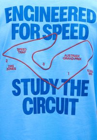 Μπλε κείμενο σε γαλάζιο ύφασμα αναγράφει "Σχεδιασμένο για ταχύτητα" και "Μελέτησε τον αγώνα" περιβάλλοντας έναν χάρτη πίστας του Γκραν Πρι Αυστρίας με κόκκινο περίγραμμα.