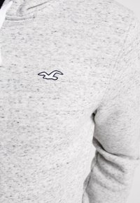 Felpa con cappuccio grigio chiaro in tessuto strutturato, con logo uccello blu navy sul petto a sinistra e cordini bianchi sul cappuccio.