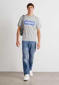 Pilkas marškinėliai su mėlynu "HUGO" logotipu, derinami su šviesiai mėlynomis džinsais ir baltais sportbačiais. Plonas, kasdieniškas dizainas su laisvu siluetu.