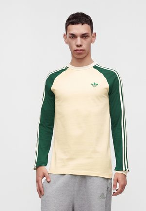 Jeune homme portant une chemise crème à manches longues avec des manches vertes et des rayures, et un pantalon de survêtement Adidas gris clair, debout devant un fond uni.