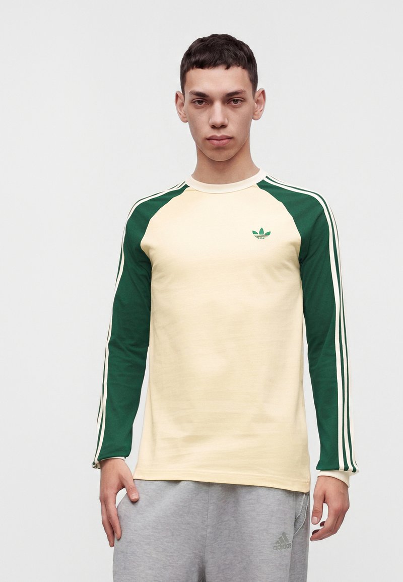 Jeune homme portant une chemise crème à manches longues avec des manches vertes et des rayures, et un pantalon de survêtement Adidas gris clair, debout devant un fond uni.