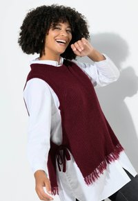 Chaleco de punto burdeos con cuello redondo, lados abiertos y flecos en el dobladillo, superpuesto a una camisa blanca con cuello.