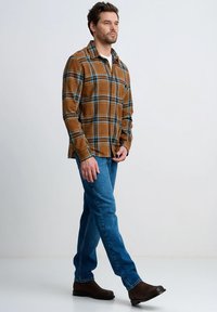 Brązowo-niebieska koszula w kratę zapinana na guziki, niebieskie dżinsy i brązowe buty. Koszula ma kieszeń i luźny krój, podkreślając wzór w kratę.
