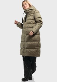 Cappotto imbottito verde oliva con cappuccio, lacci neri stringenti, zip frontale e design trapuntato. Abbinato a pantaloni neri e stivali con tacco.