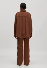 Vrouw staat met haar rug naar ons gekeerd, gekleed in een losse bruine blouse met lange mouwen en bijpassende wijde broek tegen een neutrale achtergrond.
