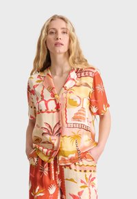 Camisa de manga corta, con botones, que presenta un vibrante estampado tropical en naranja, rosa, amarillo y crema, con un corte relajado y dos bolsillos frontales.