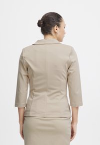 Blazer beige ajusté avec des manches trois-quarts, des coutures structurées et un col classique, confectionné en tissu lisse et extensible.