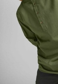 Henkilö, joka on pukeutunut oliivinvihreään pitkähihaiseen paitaan, jossa on kolme nappia kauluksessa, seisoo yksinkertaista vaaleaa taustaa vasten.