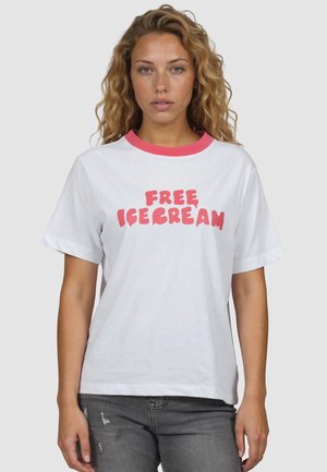 Vrouw met golvend blond haar die een wit T-shirt draagt met een roze kraag en de tekst "FREE ICE CREAM" in roze letters, gecombineerd met een grijze spijkerbroek.