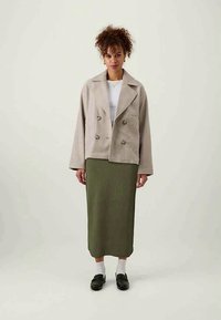 Beige, kétsoros zakó, textúrás felülettel, egy bordázott olajzöld midi szoknyával párosítva. Fekete loaferek és fehér zoknik egészítik ki a megjelenést.