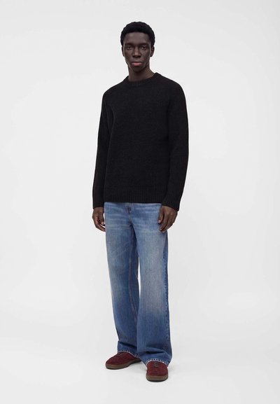 Homme debout portant un pull noir à manches longues, un jean bleu clair large et des baskets rouges sur un fond blanc uni.