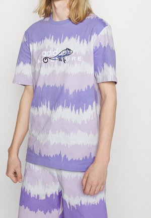 Person iført en lilla og hvid tie-dye Adidas t-shirt og matchende shorts med en lille kamæleon grafik på brystet.
