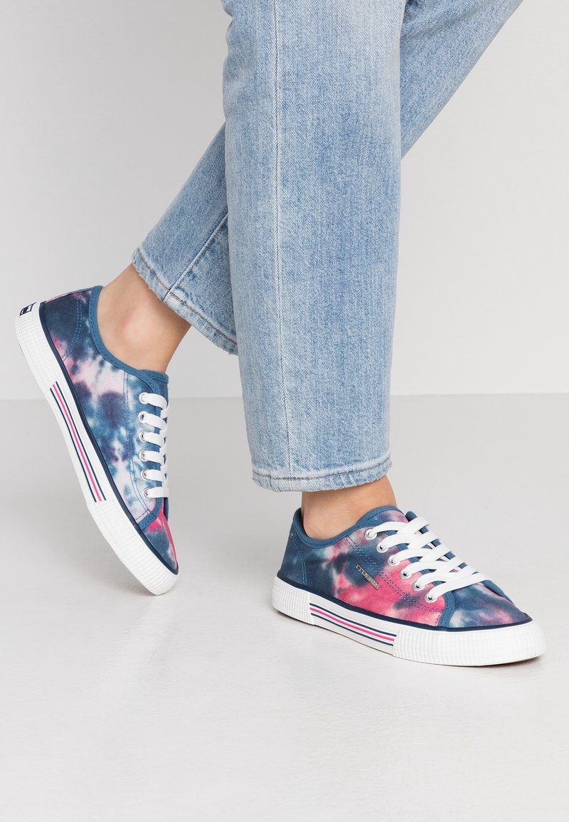 Pieds portant des baskets tie-dye bleues, roses et blanches avec des lacets blancs et des semelles rayées, assorties à un jean en denim bleu clair.