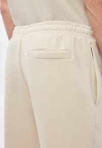 Beżowe spodnie dresowe z elastycznym pasem, posiadające płaską tylną kieszeń oraz boczne kieszenie z rozcięciami. Miękka faktura i luźny krój.