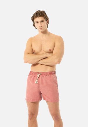 Oberkörperfreier Mann mit verschränkten Armen, der lachsfarbene Badeshorts mit weißem Kordelzug trägt und vor einem schlichten hellen Hintergrund steht.