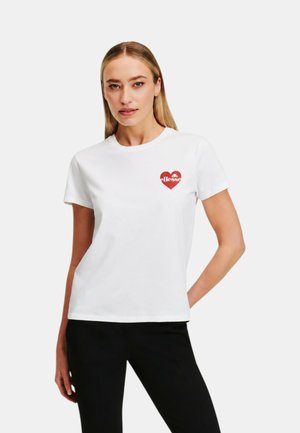 Donna con capelli biondi lisci indossa una maglietta bianca a maniche corte con un piccolo cuore rosso e un logo sul petto e pantaloni neri, in piedi con una mano sul fianco.