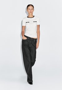 Witte t-shirt met korte mouwen en zwarte afwerking, voorzien van twee voorkruiden; gecombineerd met losse, donkere denim jeans; model staat met handen in de zakken.