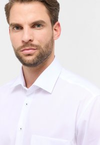 Camicia bianca con colletto classico, texture delicata e chiusura con bottoni. Include una singola tasca sul petto e dettagli in bottoni lucidi.
