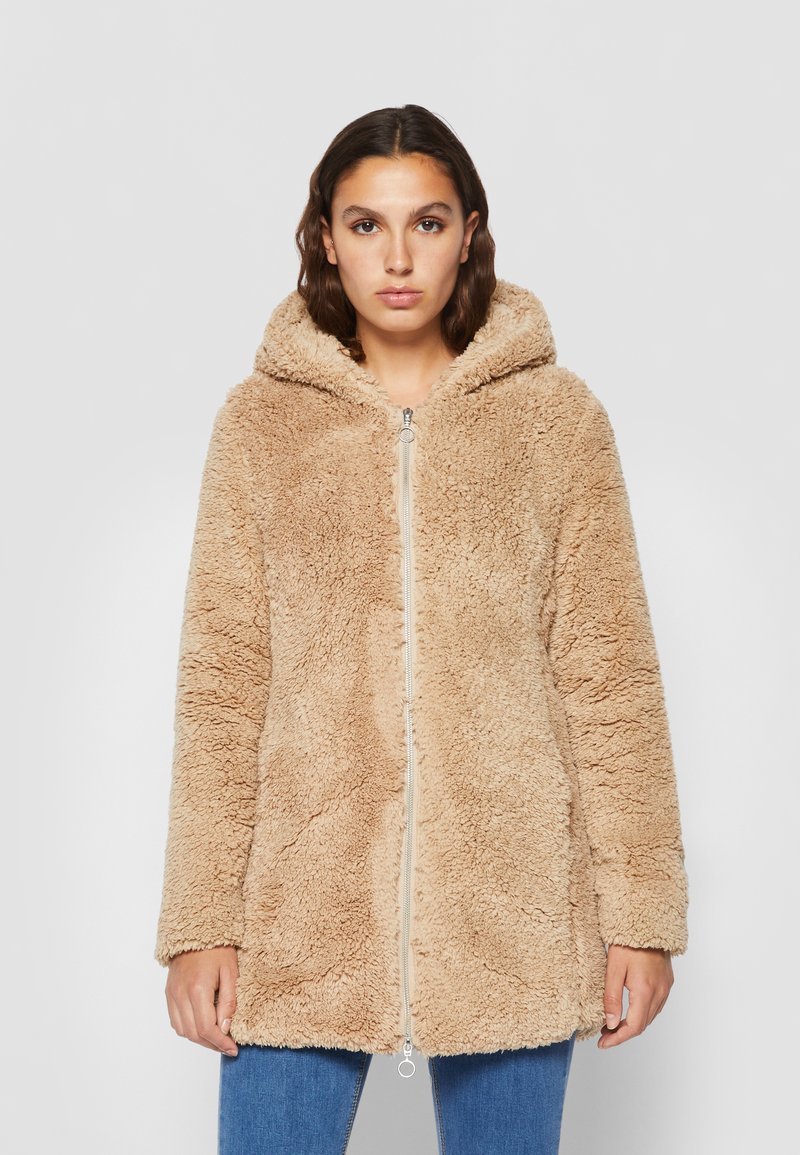 Beige, pörröinen tekoturkki takki hupulla, edessä vetoketju ja pitkät hihat, joissa on pehmeä rakenne ja rento istuvuus.