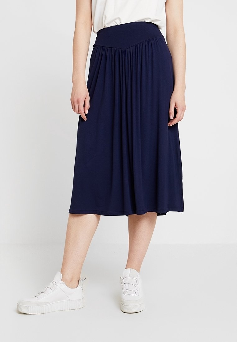 Femme portant une jupe midi bleu marine avec des détails froncés, associée à des baskets blanches et un haut blanc, se tenant devant un fond uni.