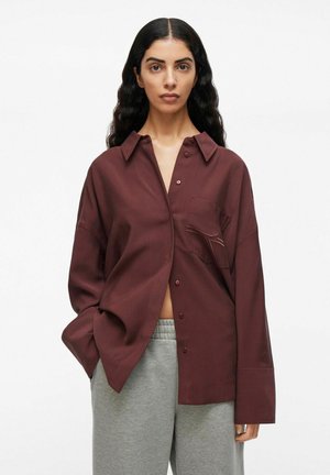 Bordeaux oversized shirt met een puntige kraag, voorzien van een enkele borstzak en contrasterende stiksels, gecombineerd met grijze sweatpants.