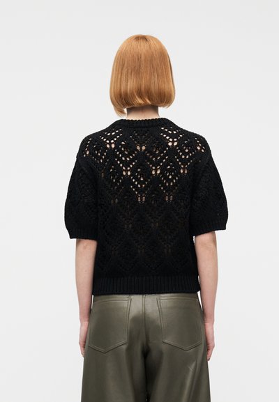 Femeie cu păr roșu scurt, purtând un pulover negru cu mâneci scurte și tricotaj deschis și pantaloni de piele verde măsliniu, văzută din spate.