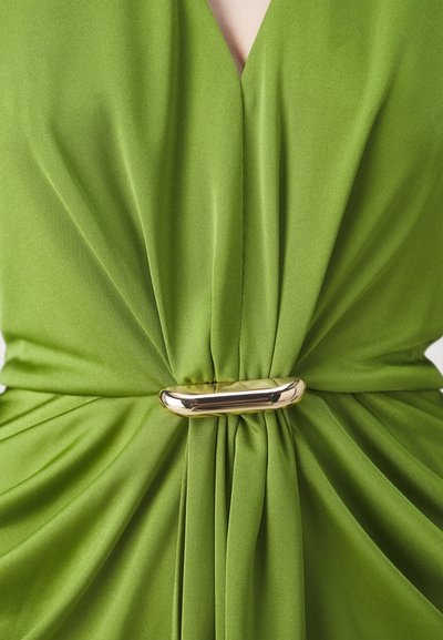 Rochie verde cu țesătură moale și texturată, având un design drapat. Accesoriul din aur de la talie accentuează aspectul strâns.