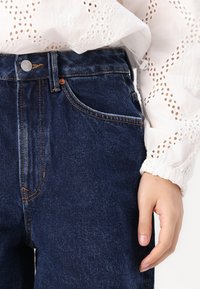 Mörkblå denimjeans med en klassisk passform, med orange sömmar och en framficka, tillsammans med en vit blus med hålmönster.