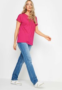 Top fucsia a maniche corte con scollo a V e dettagli in bottone, abbinato a jeans blu chiaro e sneakers bianche con accenti grigi.
