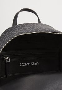 Czarny plecak z wzorzystą zewnętrzną powłoką, srebrnym zamkiem błyskawicznym i skórzanym wnętrzem. Zawiera kieszeń na zamek i tłoczoną nazwę marki wewnątrz.