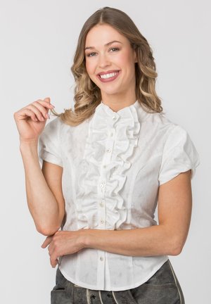 Lächelnde Frau mit welligem blondem Haar, die eine weiße Rüschenbluse und dunkle Hosen trägt, posiert mit einer Hand erhoben und der anderen verschränkt.