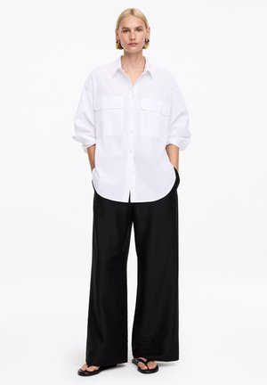 Mujer de pie mirando hacia adelante, vestida con una camisa blanca de botones de gran tamaño, pantalones anchos negros y sandalias negras tipo chancla sobre fondo blanco.
