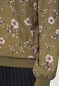 Oliven grøn blomstret bluse med pink blomster, der har ribbede manchetter og en sidelomme. Glat tekstur og afslappet pasform.