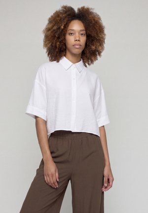 Mujer con pelo rizado que lleva una camisa blanca de manga corta y botones, suelta, y pantalones marrones de cintura alta y pernera ancha, de pie frente a un fondo sencillo.