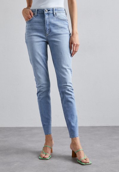 Jeanși skinny de culoare albastru deschis, din denim, cu talie înaltă, buzunare frontale și tiv brut la glezne. Asortați cu sandale verzi cu curele.