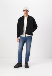 Jachetă bomber neagră cu fermoar frontal, manșete cu nervuri și un logo subtil; asortată cu pantaloni lejeri din denim deschis și tenisi închiși la culoare.