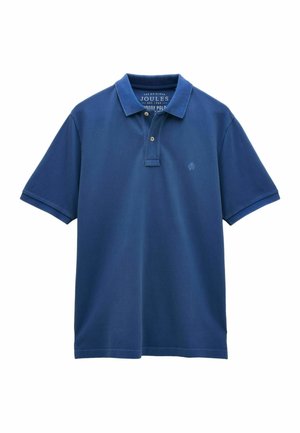 Man wearing blue polo shirt, beige shorts, and patterned slip-on shoes standing against a plain beige wall.